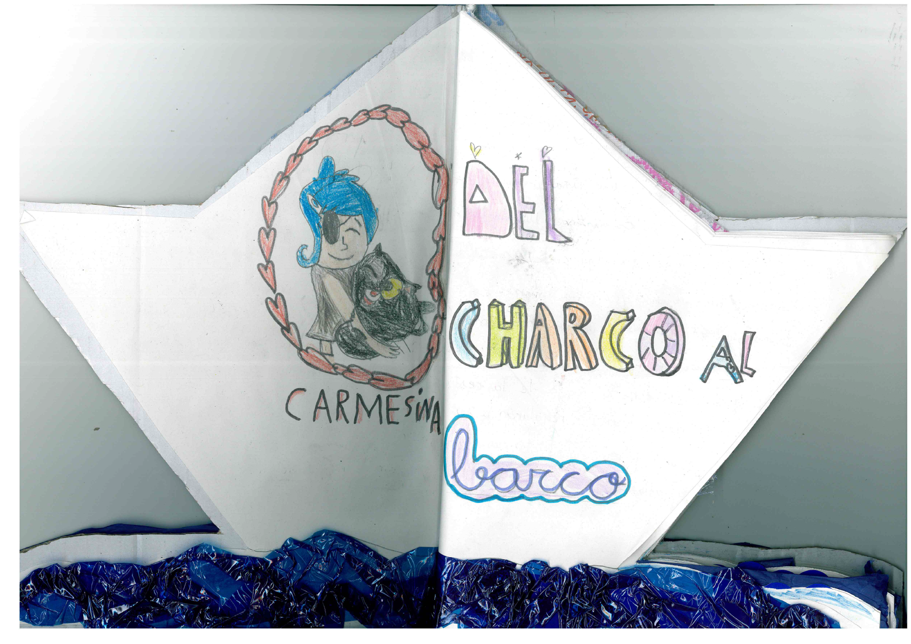 Imagen de un barco recortado de un folio en el que aparece el t tulo del libro, construido con cart n, y en la parte de la izquierda hay un dibujo infantil de Carmesina, con el pelo azul, un parche en el ojo y sujetando lo que parece ser un animal de color negro, con grandes ojos. En la parte de abajo se aprecia que en la base del barco hay papel charol de color azul simulando las olas del mar. Todas las p ginas est n escritas en un papel recortado simulando el contorno de un barco.